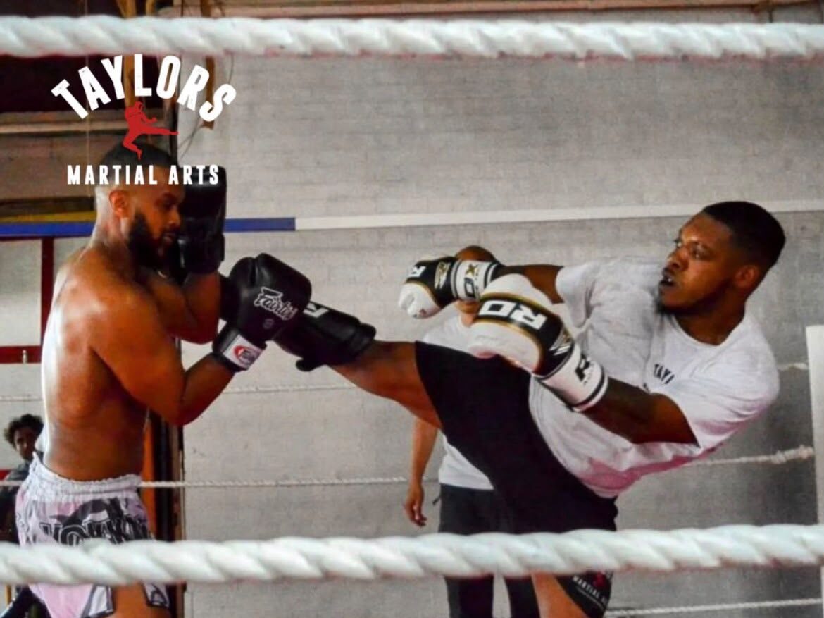 Adult freestyle kickboxing at Taylor's Martial Arts Two adult students sparring in the ring at Taylor's Martial Arts, one landing a high kick to the guard