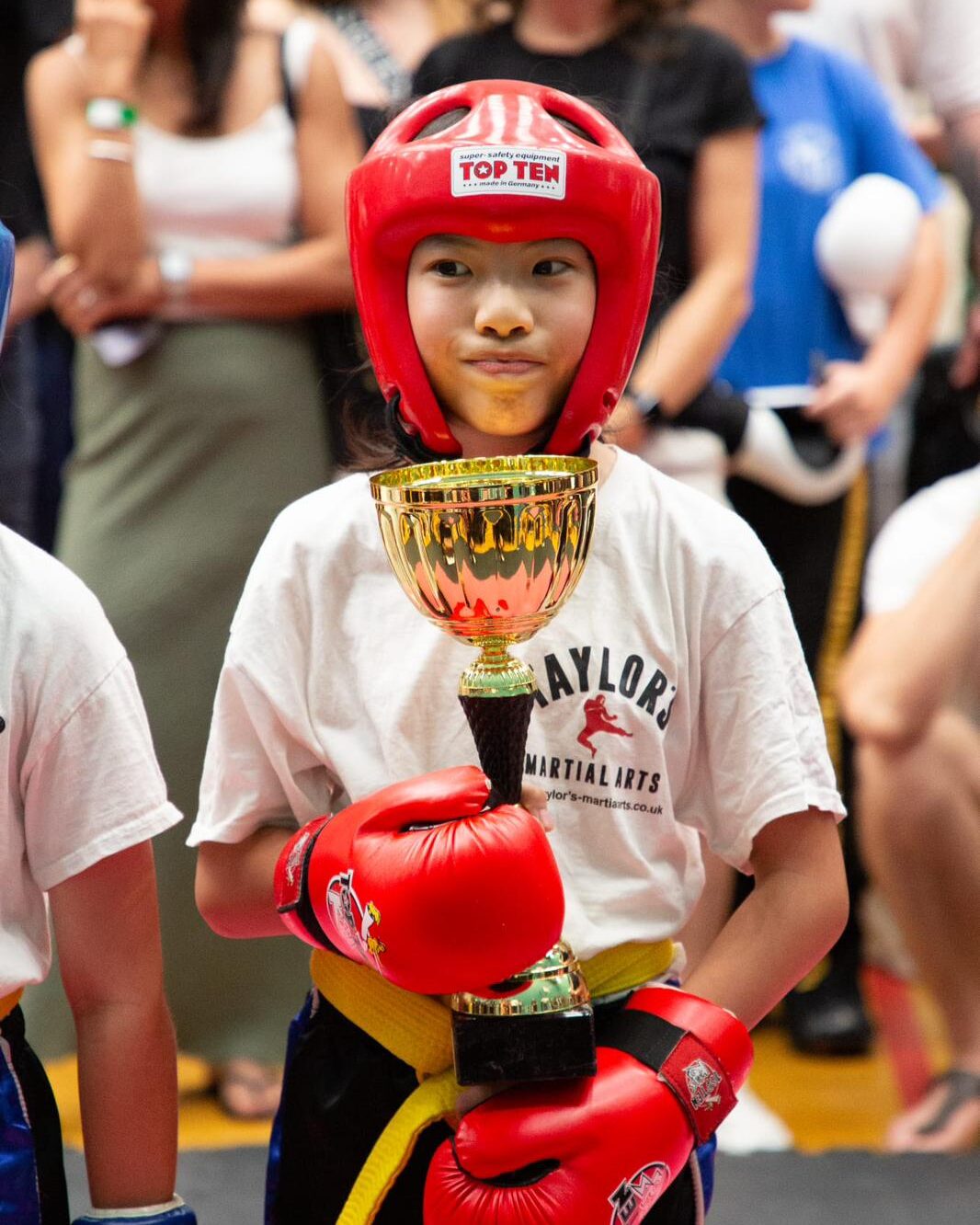 A young Taylor's Martial Arts student in a red headguard holding a gold competition trophy