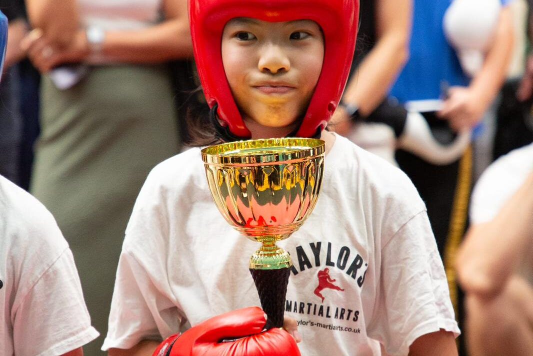 A young Taylor's Martial Arts student in a red headguard holding a gold competition trophy